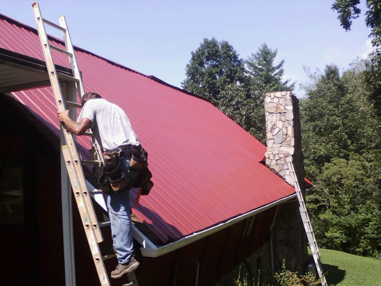 Roofer on a ladder beside a red metal roof and stone chimney — Soffit & Fascia Repair by Roofing Brothers in Nazareth